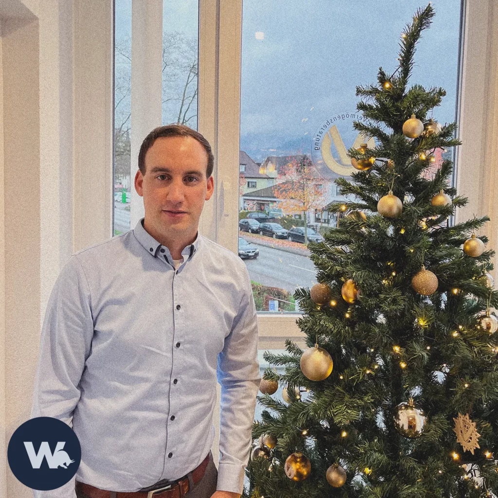 Simon steht neben einem Weihnachtsbaum, verziert mit goldenemS Schmuck. Im Hintergrund sieht man den Ausblick seines Büros in Waldkirch.